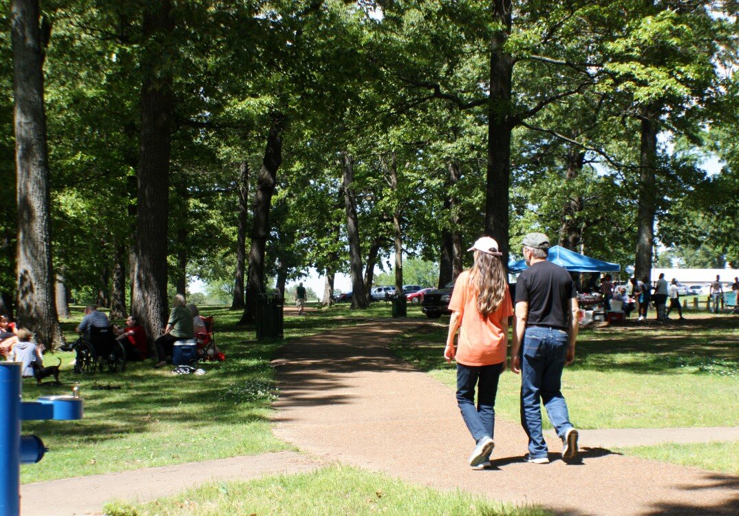 Park goers walk the path at Audubon Park between a picnic to the left and cookout to the right. Classic soul music plays from a second cookout, which they've just passed. April 2021. (Cole Bradley) 