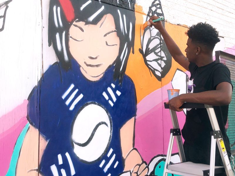 Donte Davis works on a section of the You's Grocery mural depicting store owner Ae An Sartain as a child when her father first opened the store. The artists based the mural on Sartain's memories and neighborhood history. (A.J. Dugger)