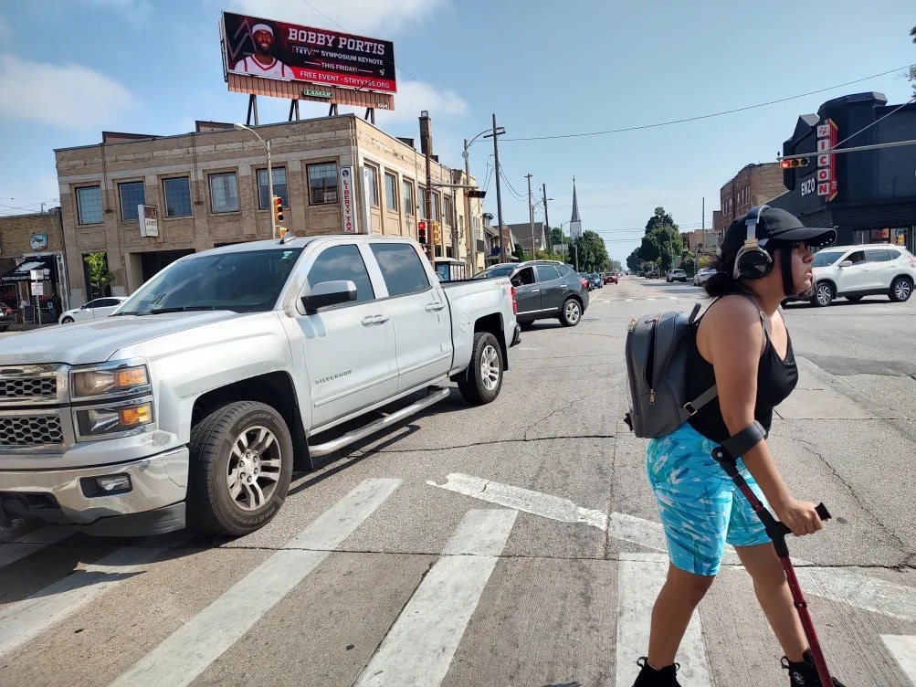 A young woman using a cane walks across the street using a cross walk.